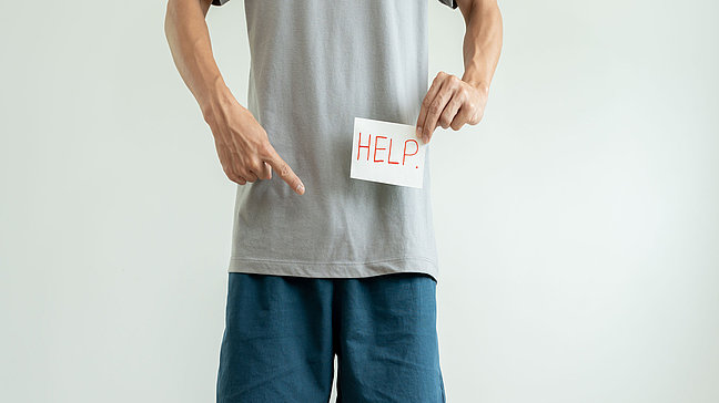 Penisschmerzen - Hinweis auf Schmerzen und Hilfebedarf. Urologenzentrum Wien - Penisschmerzen: Mann zeigt auf ein Schild mit der Aufschrift "HELP" vor seinem Unterkörper.