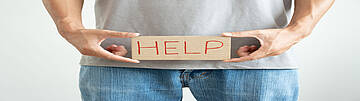 Hypospadie - Symbolische Darstellung von Hilfebedarf. Urologenzentrum Wien - Hypospadie: Mann hält ein Schild mit der Aufschrift "HELP" vor seinem Unterkörper.
