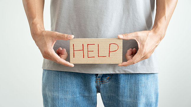 Hypospadie - Symbolische Darstellung von Hilfebedarf. Urologenzentrum Wien - Hypospadie: Mann hält ein Schild mit der Aufschrift "HELP" vor seinem Unterkörper.